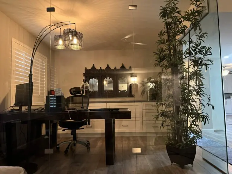 Modern home office with glass partition, black desk, computer monitor, arc lamp with hanging pendant lights, decorative mirror, potted bamboo plant, and wooden flooring