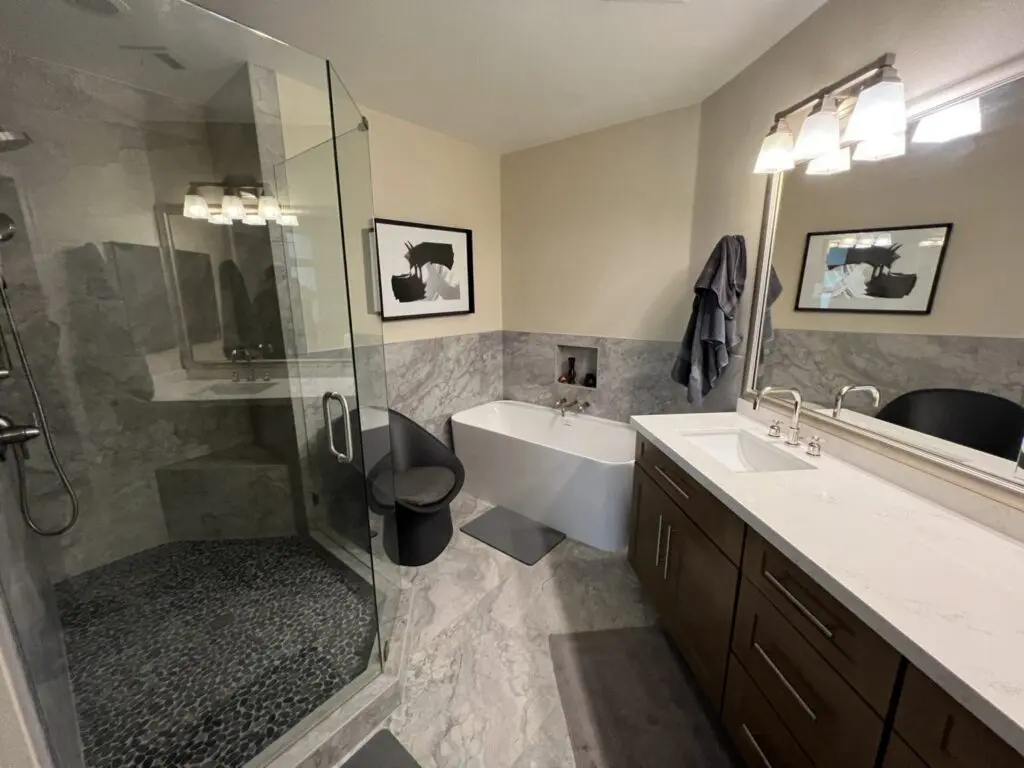Modern master bathroom with gray marble tile, walk-in shower with glass enclosure, freestanding white soaking tub, dual vanity sink with brown cabinetry, and framed artwork above