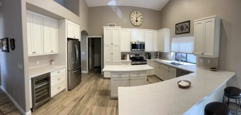 Modern kitchen with white cabinetry, stainless steel appliances, large island with white countertop, wood-grain flooring, and neutral beige walls with a clock