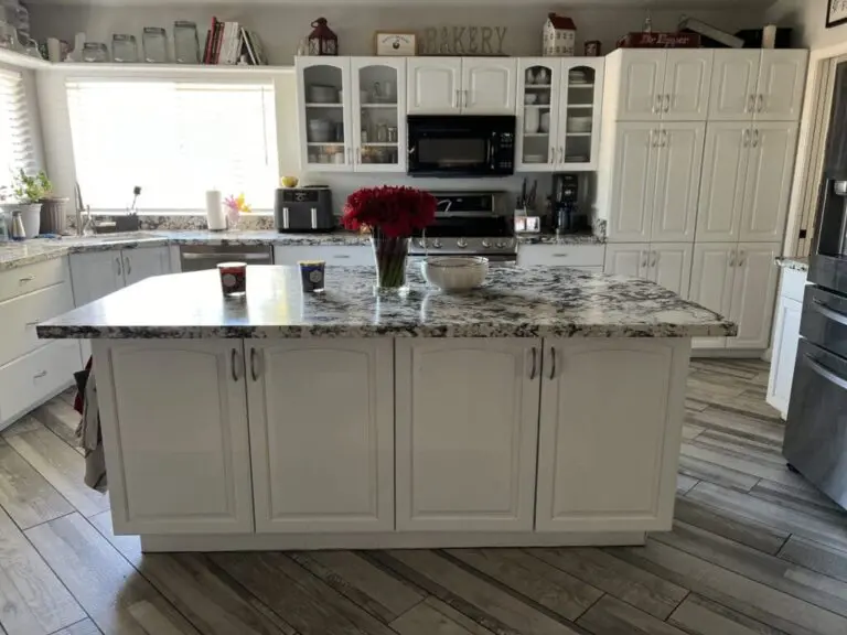 Modern kitchen with cream cabinetry, granite countertops, large island with seating, stainless steel appliances, and wood-look flooring with red flowers in vase