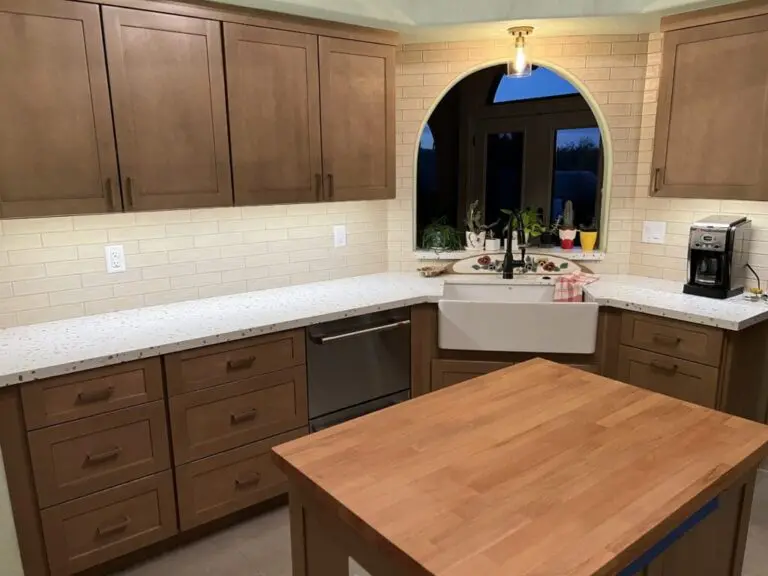 Modern kitchen with brown cabinetry, white quartz countertops, farmhouse sink, subway tile backsplash, and arched window overlooking plants and evening sky