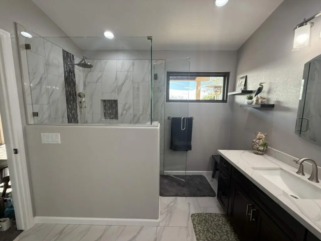 Modern bathroom with walk-in shower, marble tile, dark vanity with white countertop, dual sinks, and floating shelves near window