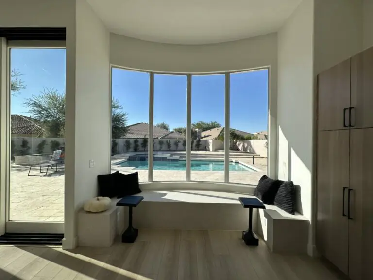 Modern home interior with curved bay windows overlooking a swimming pool and patio, featuring built-in window seating with black pillows and dark wood cabinetry
