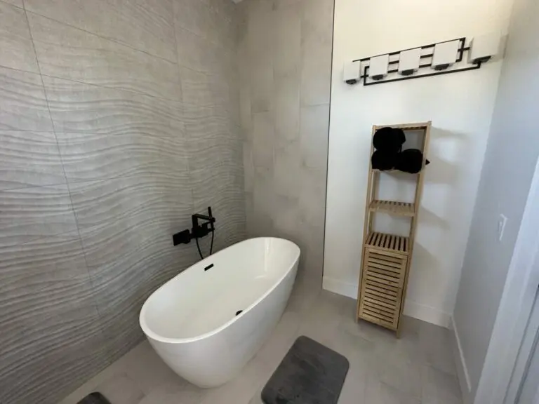 Modern bathroom with freestanding white soaking tub, gray wave-pattern tile surround, wooden towel ladder, and black bath mat