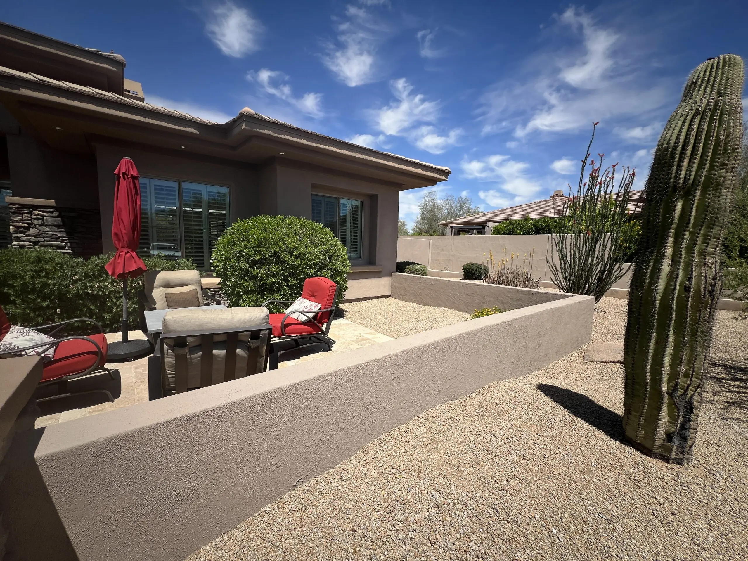 Modern desert patio with lounge chairs, red umbrellas, and tall saguaro cactus near contemporary home with stone walls and gravel landscaping