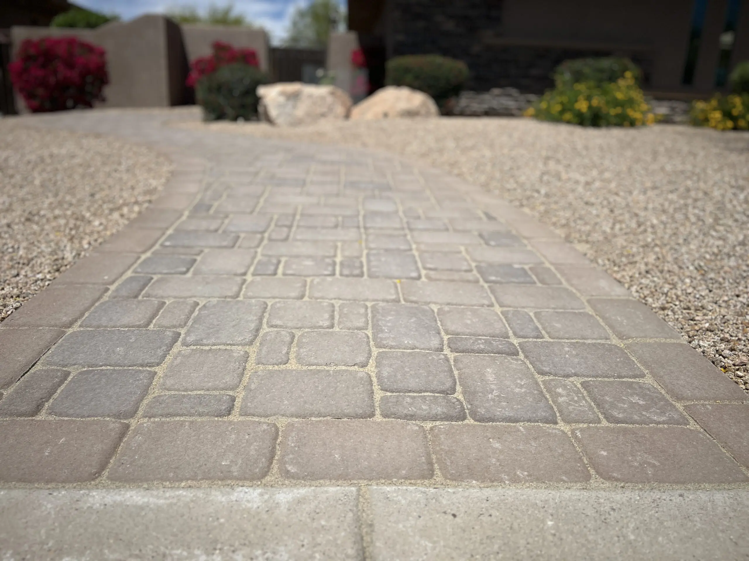 Low-angle view of a geometric patterned paver pathway in a landscaped cemetery or memorial garden with flowering plants and stone monuments in the background