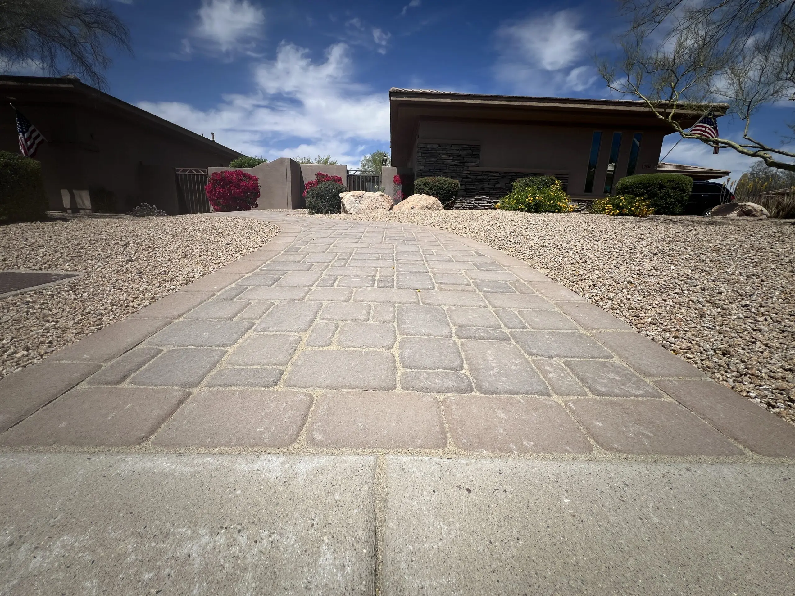 Paved driveway leading to modern desert-style home with brown exterior, surrounded by gravel landscaping and flowering shrubs under blue sky