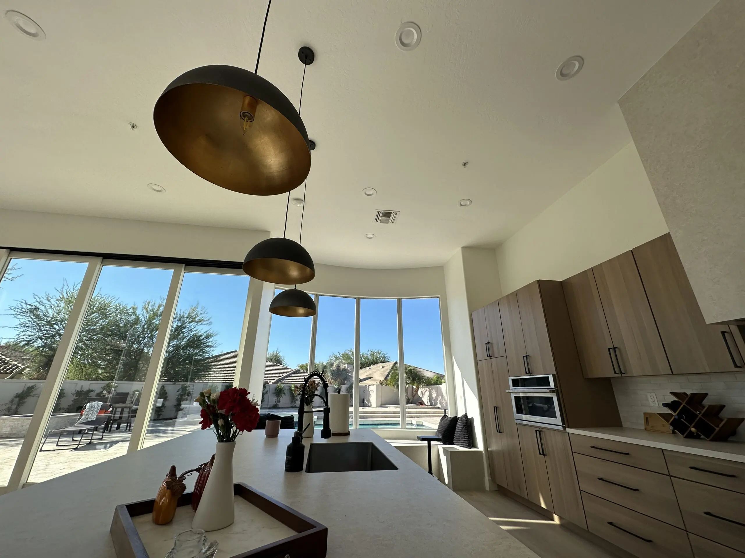Modern kitchen with three brass pendant lights hanging above white quartz island, floor-to-ceiling windows overlooking backyard, light wood cabinetry, and stainless steel appliances