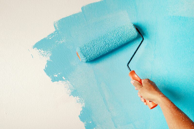 A hand holds a paint roller, applying a fresh coat of blue paint to a wall, signifying the beginning stages of a kitchen remodeling project.