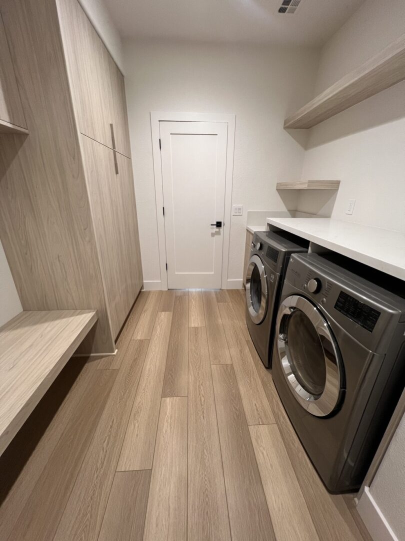 Modern laundry room with two front-load washers, white countertop, wood-grain cabinetry, floating shelves, and light wood plank flooring