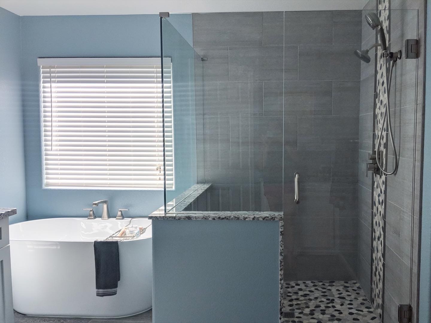 Modern bathroom with freestanding white soaking tub, large walk-in shower with gray tile and black pebble floor, blue walls, and white blinds covering window