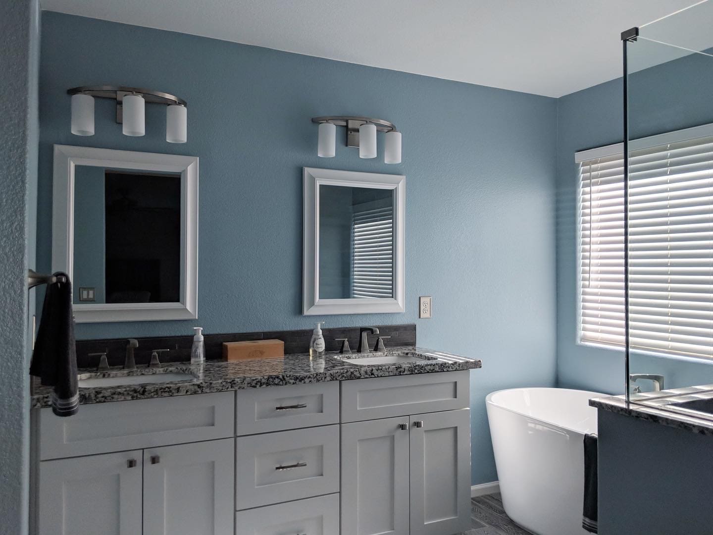Modern bathroom with light blue walls, gray vanity cabinets, granite countertop, dual framed mirrors with sconces, freestanding white tub, and glass shower enclosure with shuttered window