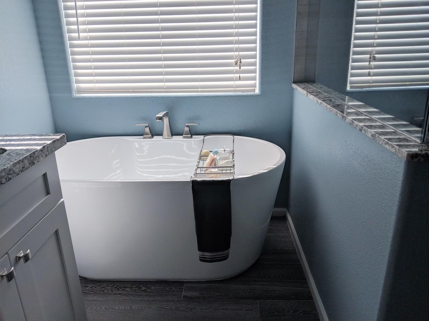 Modern bathroom with freestanding white soaking tub, chrome faucet, gray granite countertops, white vanity, and horizontal blinds on windows