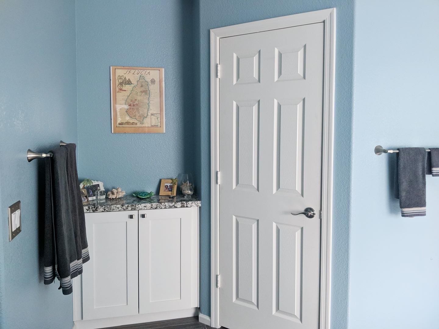 Light blue bathroom with white six-panel door, white vanity cabinet with granite countertop, dark gray towels on brass racks, framed map of St. Lucia on wall, and decorative items on counter