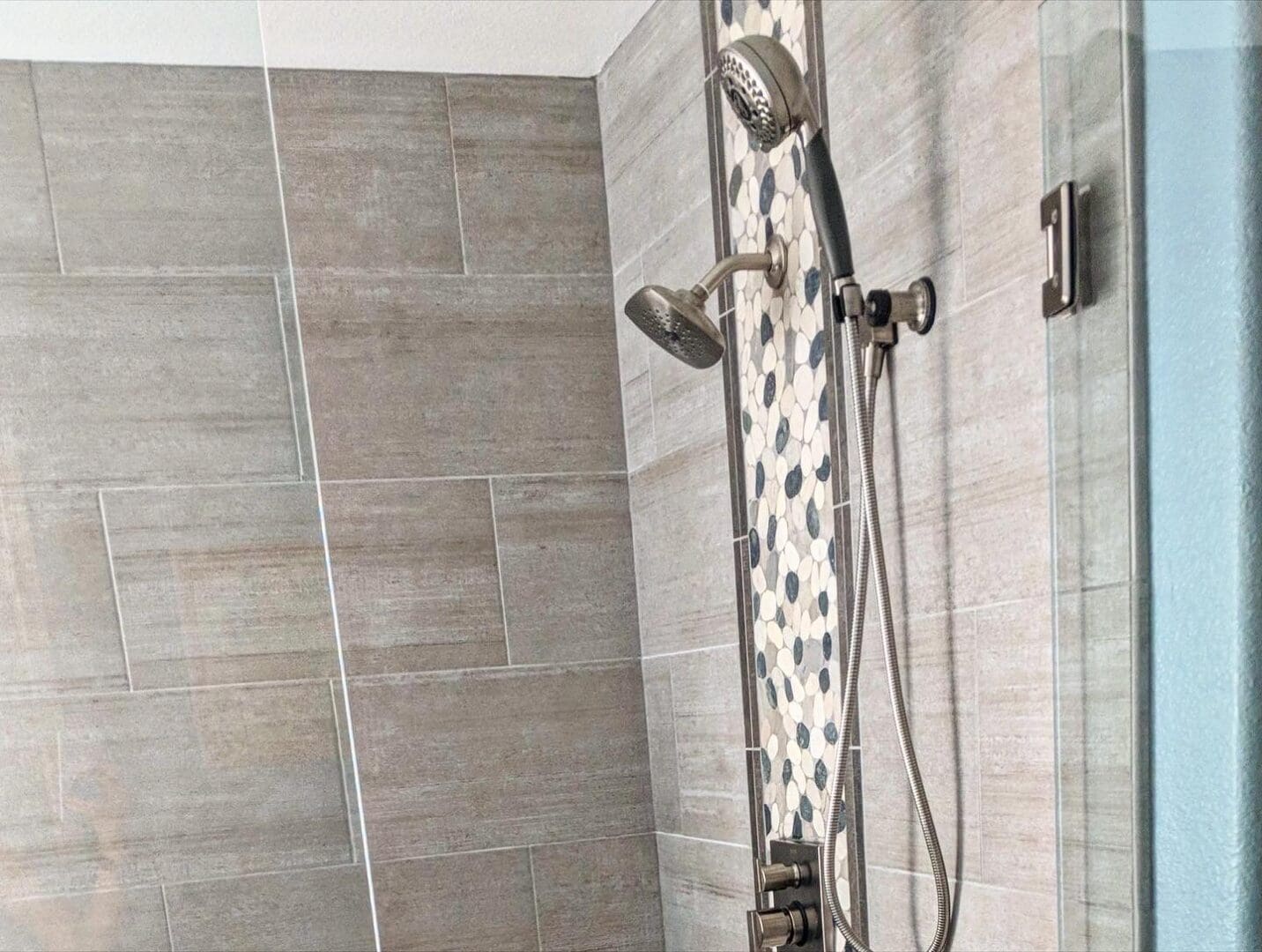Modern shower enclosure with dual showerheads, gray wood-grain tiles, and white pebble accent strip against glass door