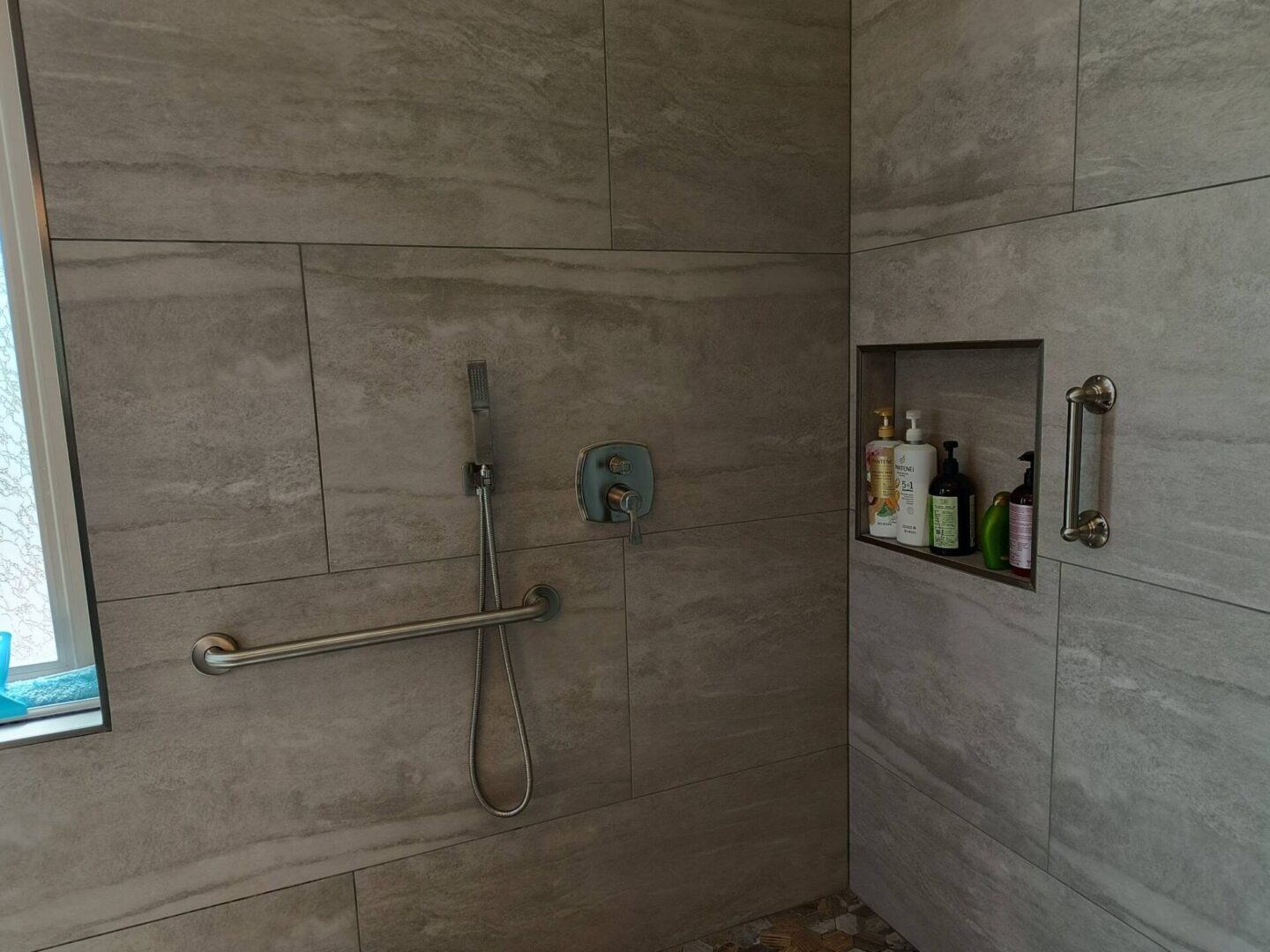 Modern walk-in shower with gray tile walls, brushed nickel fixtures, grab bar, handheld showerhead, and recessed shelving holding toiletries