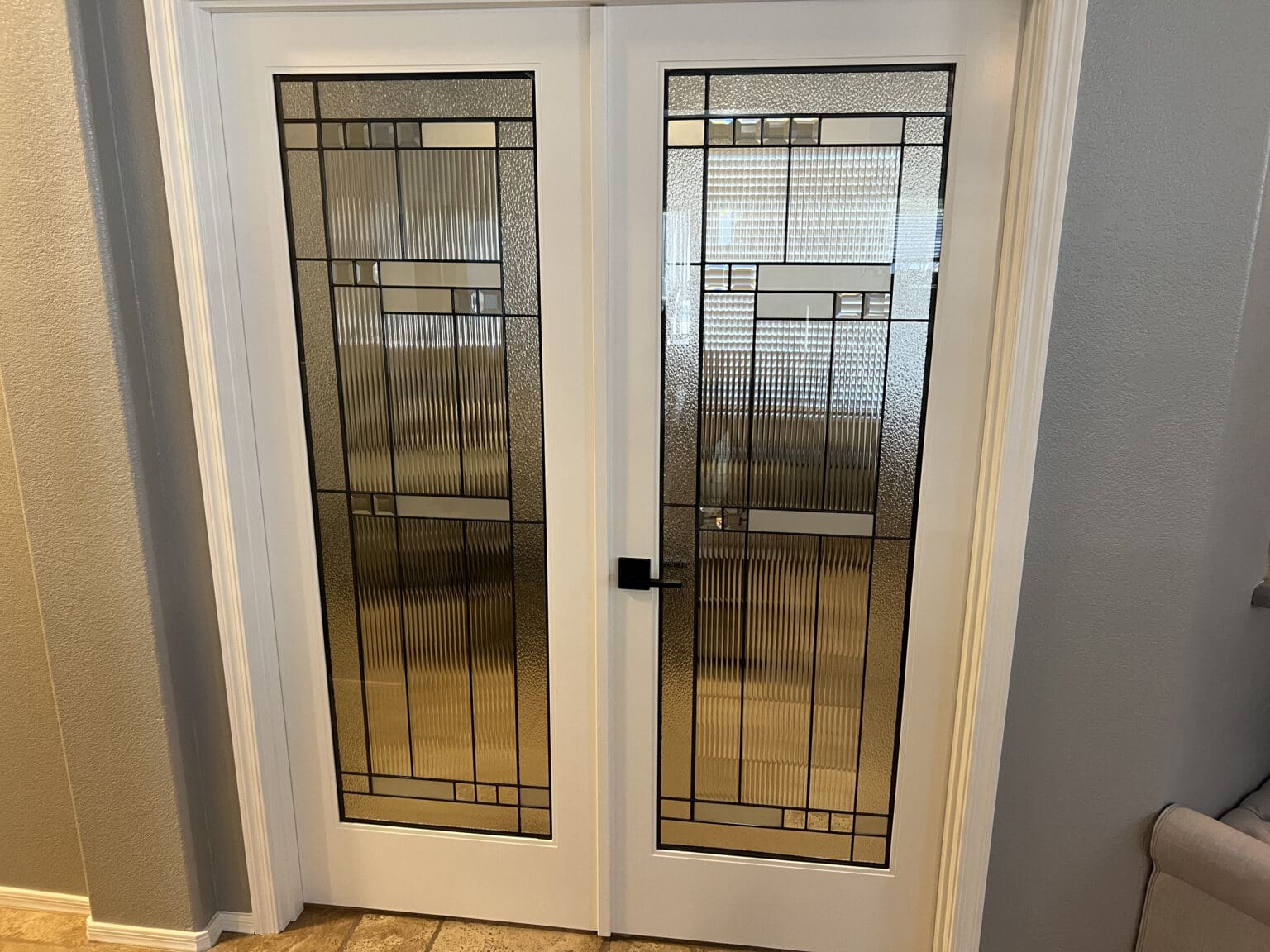 Two white French doors with black-framed geometric glass panels featuring vertical ribbed texture, located in a modern interior space with cream-colored walls