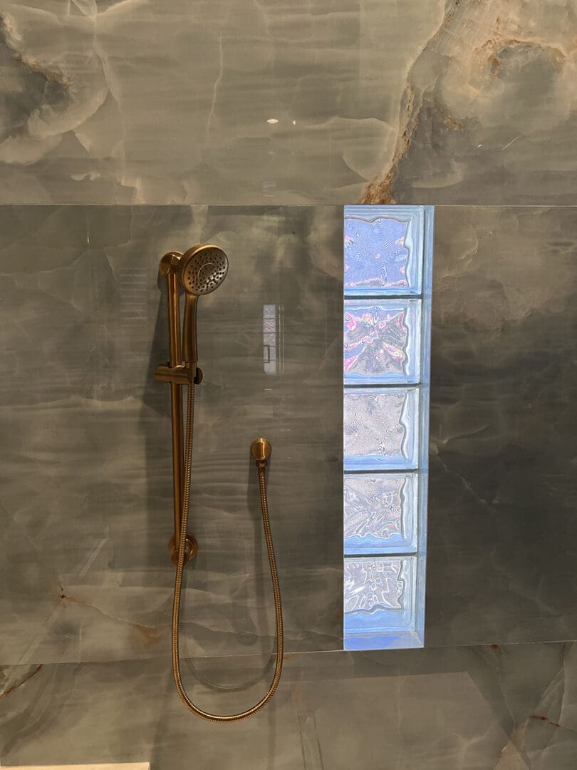 Bathroom shower display featuring a bronze handheld showerhead with hose on a gray tile wall, alongside decorative glass block accents in blue and white tones