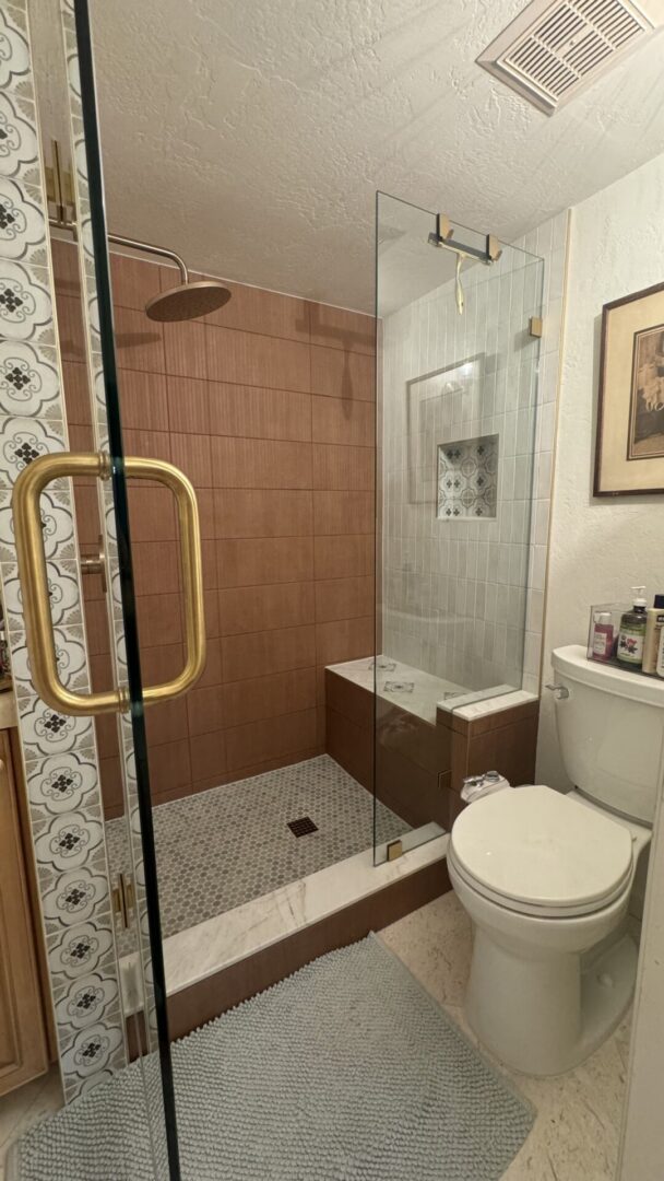 Modern bathroom with walk-in shower featuring brown tile walls, glass enclosure with gold handle, rainfall showerhead, and white subway tile accents, adjacent to white toilet and decorative patterned wallpaper
