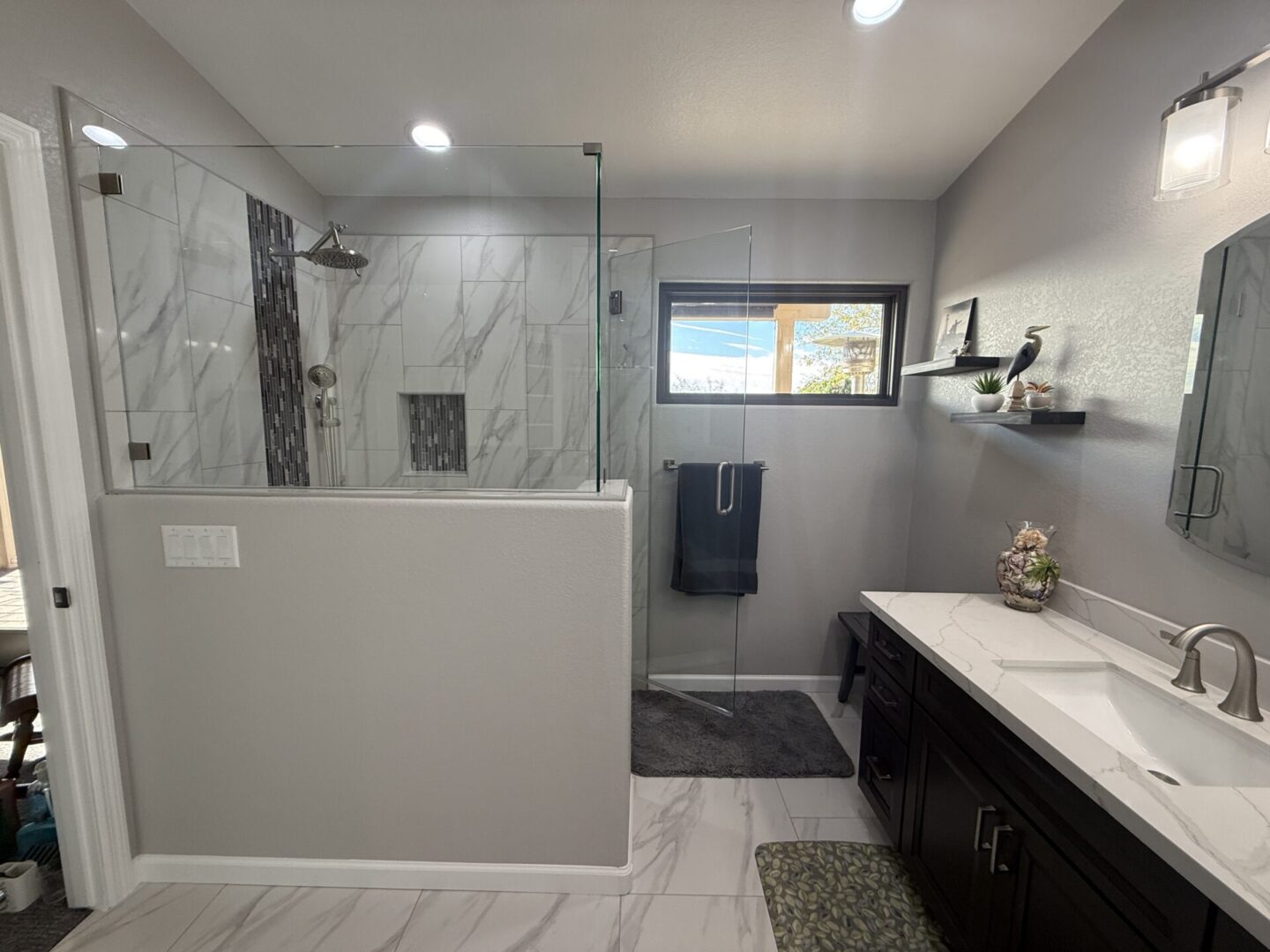 Modern bathroom with walk-in shower, marble tile, dark vanity with white countertop, dual sinks, and floating shelves near window