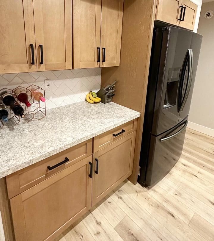 Modern kitchen with natural wood cabinets, white subway tile backsplash, speckled gray countertop, black stainless steel refrigerator, and light wood flooring