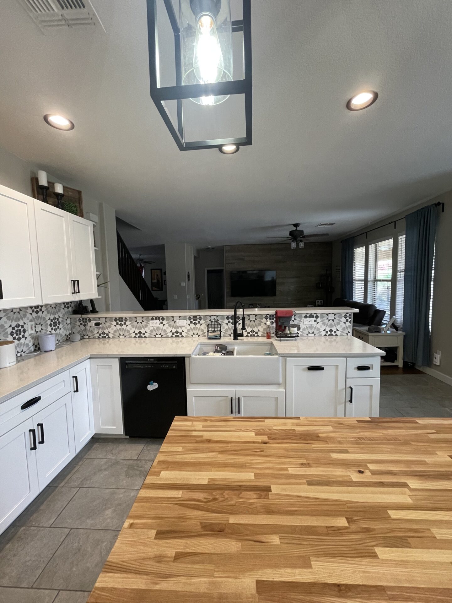 Modern kitchen with white cabinetry, farmhouse sink, black appliances, patterned backsplash, and wooden island countertop overlooking open living area with dark wood accent wall
