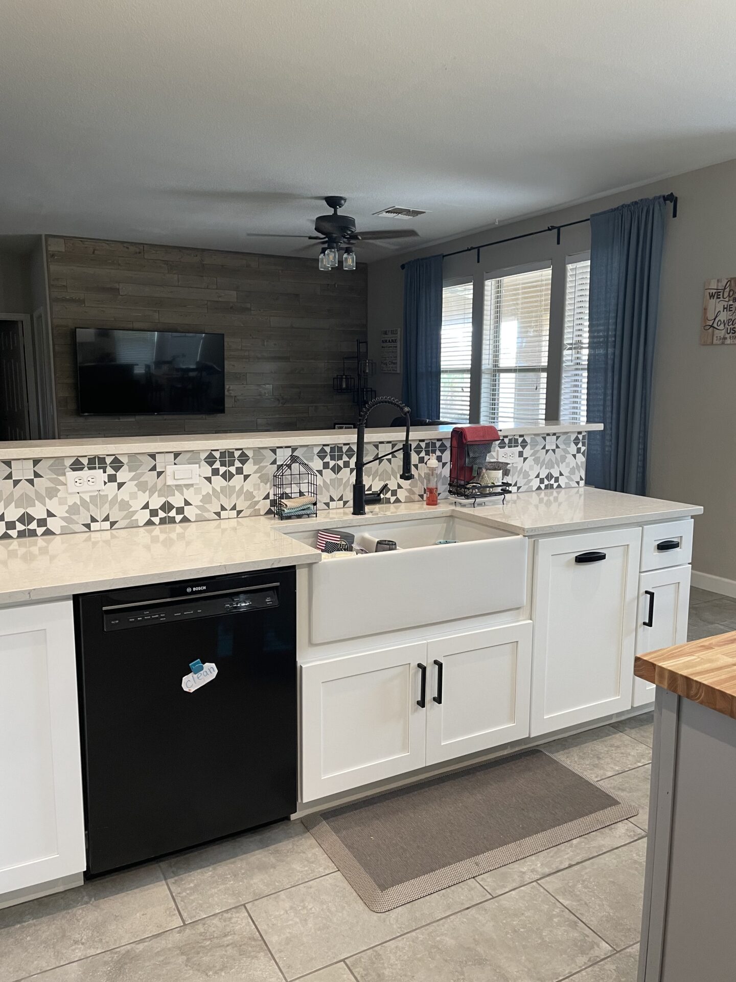 Modern kitchen with white cabinetry, farmhouse sink, black dishwasher, geometric tile backsplash, quartz countertops, blue curtains, and ceiling fan overlooking open concept living area