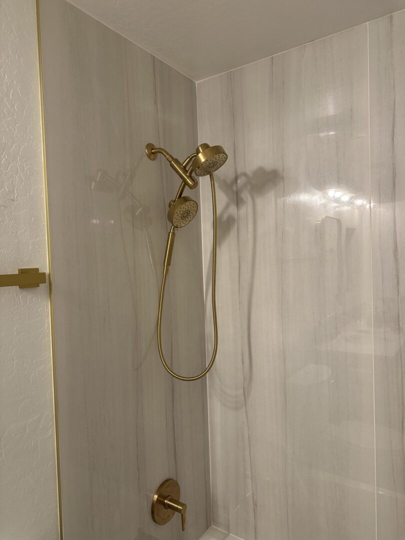 Modern bathroom shower enclosure with dual brass showerheads and handheld spray on light marble tile walls with gold trim