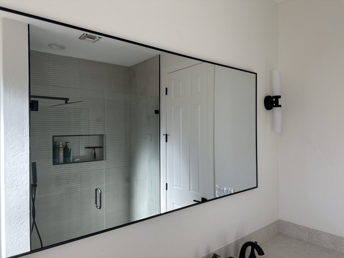 Modern bathroom with large framed mirror above counter, glass-enclosed shower with textured tile walls and built-in shelf, black hardware and fixtures