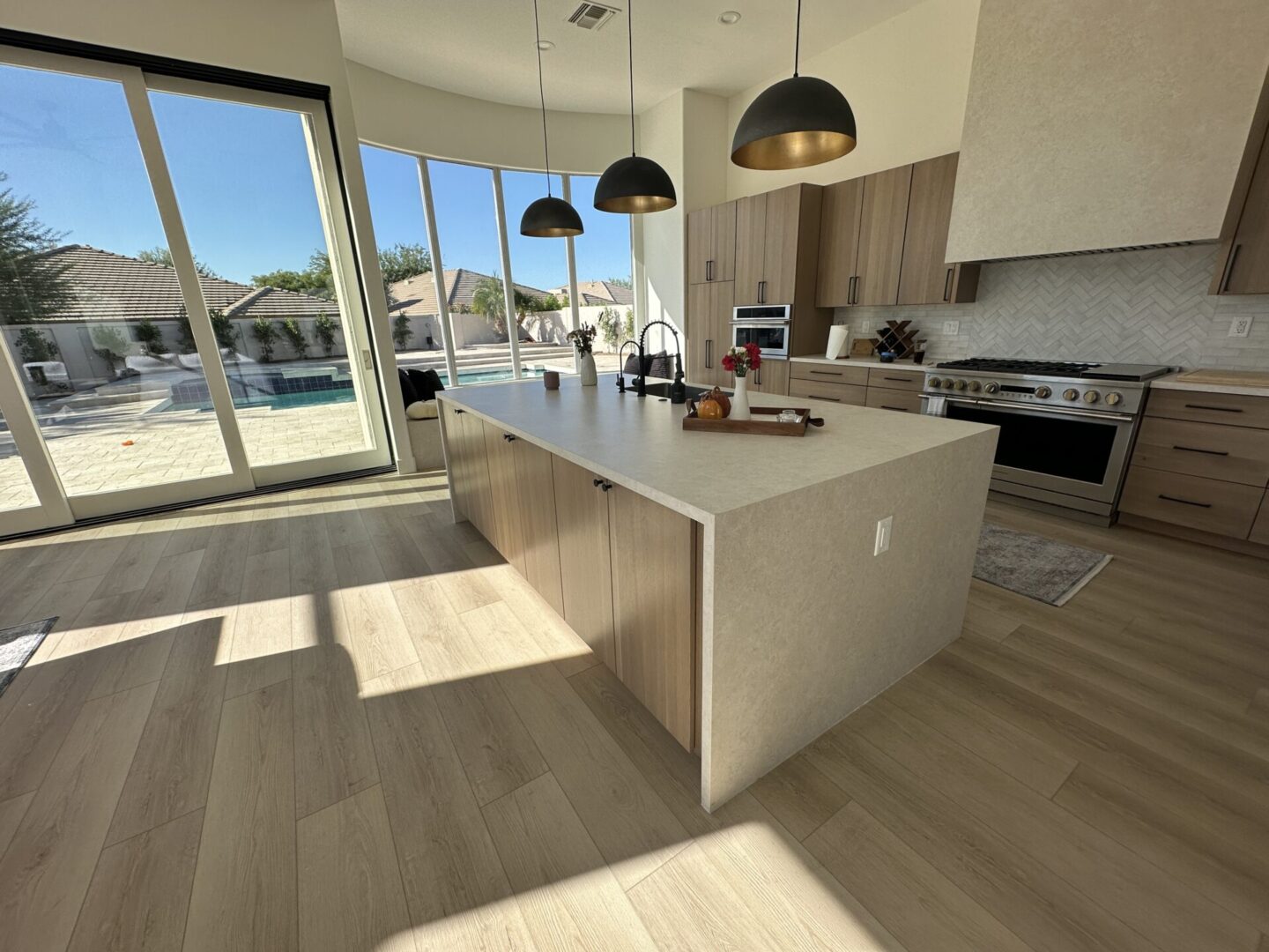 Modern luxury kitchen with large island, pendant lights, and floor-to-ceiling windows overlooking a swimming pool and residential neighborhood
