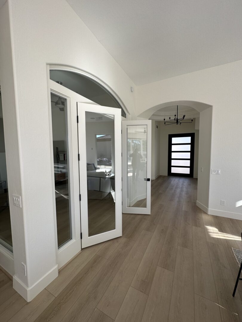 Modern hallway with white walls, wood-look flooring, arched doorways, glass-panel doors leading to living spaces, and a contemporary black front door with horizontal window panels at the end
