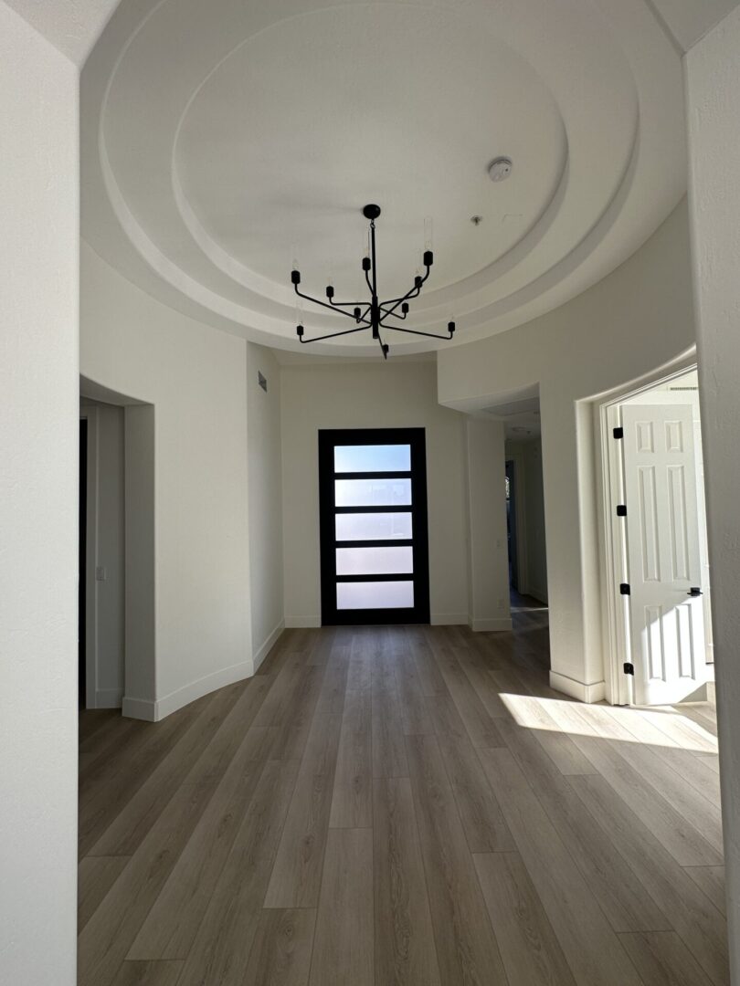 Modern foyer with curved coffered ceiling, black chandelier, wood plank flooring, black contemporary front door with horizontal glass panels, white interior doors, and natural light from right