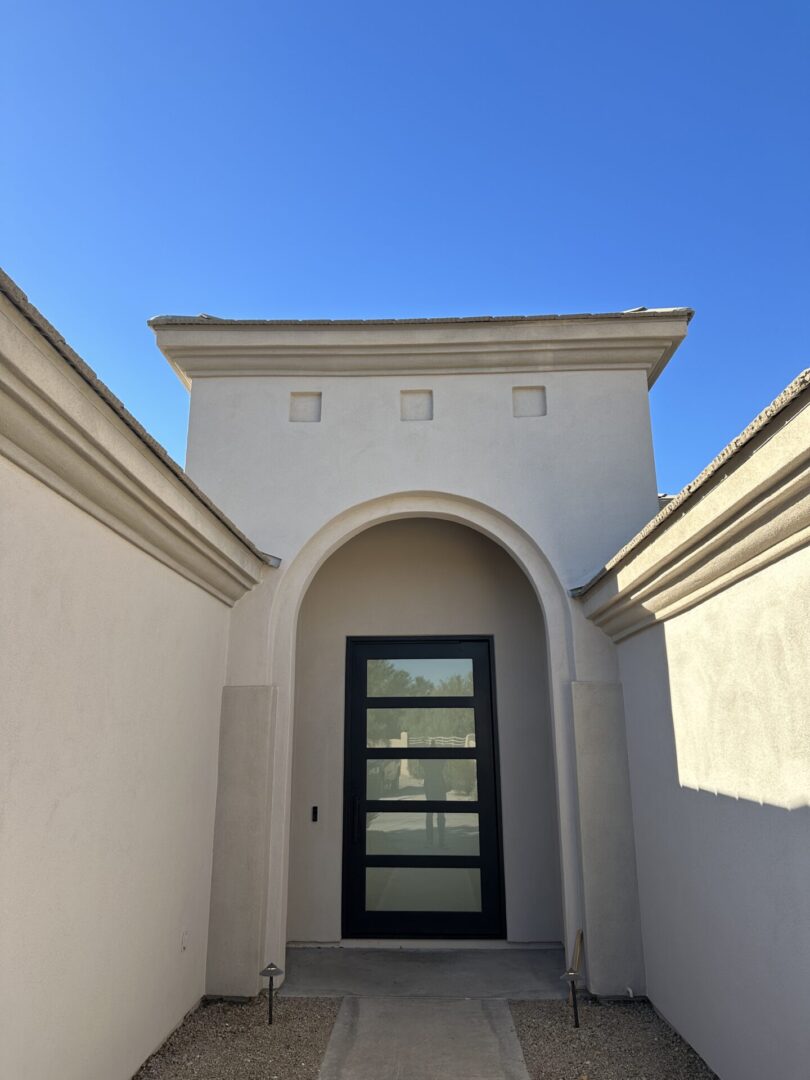 Modern white building entrance with black glass door featuring five horizontal panes, flanked by curved archway and symmetrical white walls under clear blue sky