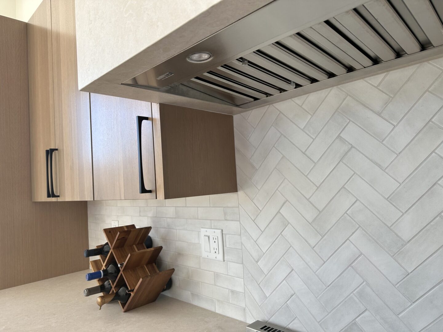 Modern kitchen with light wood cabinets, stainless steel range hood, white subway tile backsplash in herringbone pattern, wooden wine rack, and electrical outlet on tiled wall