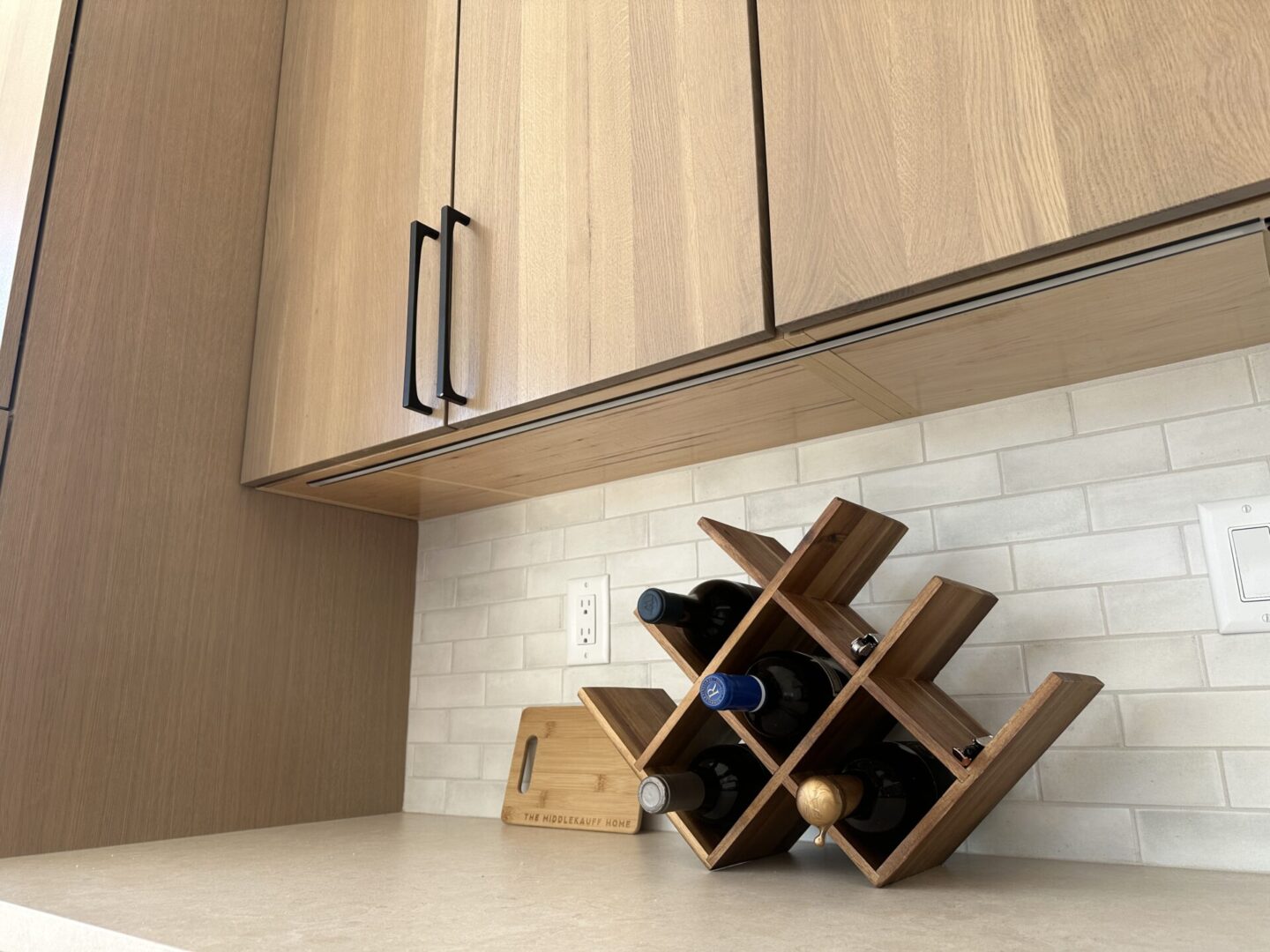 Modern kitchen countertop with wood-grain upper cabinets, white subway tile backsplash, and geometric wooden wine rack holding multiple bottles