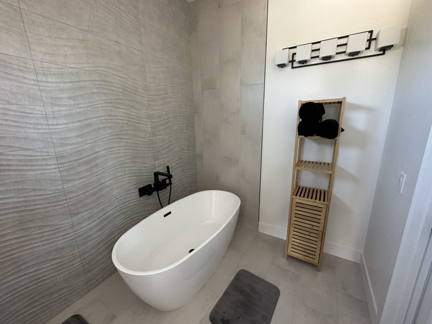 Modern bathroom with freestanding white soaking tub, gray wave-pattern tile surround, wooden towel ladder, and black bath mat