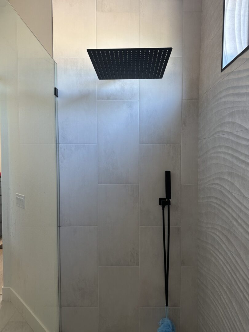 Modern bathroom shower with large format gray tile walls, black rectangular rainfall showerhead mounted on ceiling, and black handheld shower wand with hose against textured accent wall