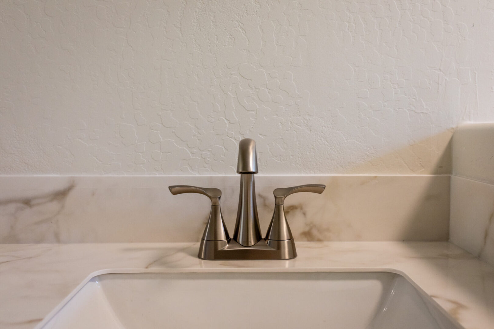 Brushed nickel two-handle bathroom faucet mounted on white marble countertop above ceramic sink with textured wall backsplash