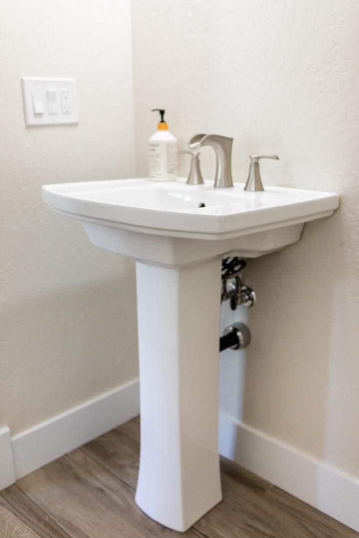 White pedestal sink with chrome faucet and soap dispenser in a modern bathroom with beige walls and wood-look flooring