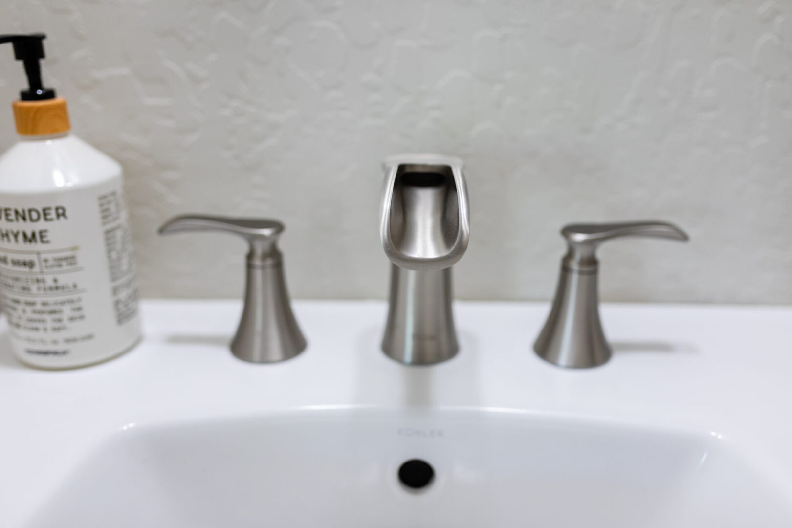 Modern bathroom sink with three brushed nickel faucet handles and a white soap dispenser with bamboo pump top