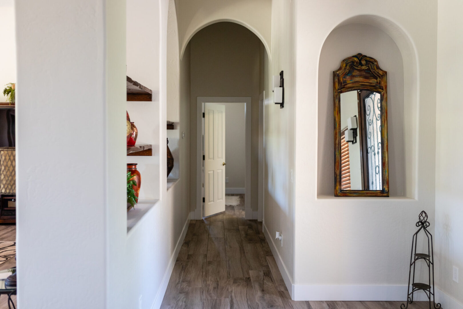 Bright hallway with arched doorways, decorative wooden mirror in alcove, dark wood flooring, and shelving with ornamental items on white walls