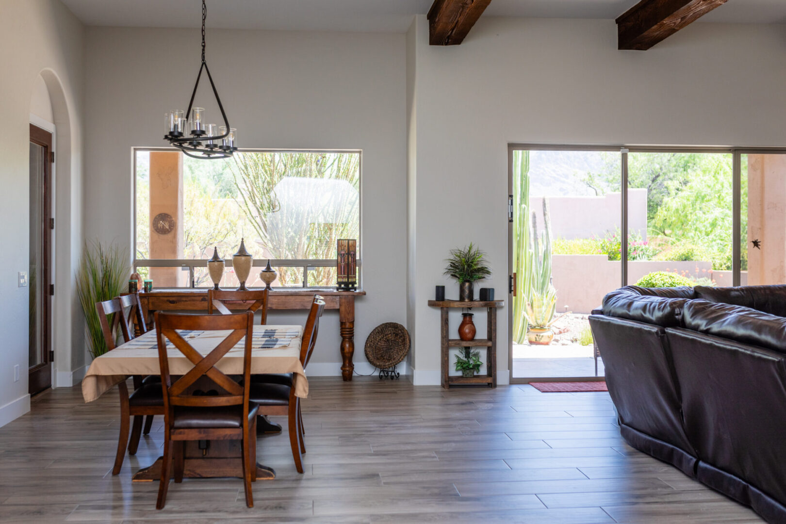 Modern open-concept living room with dining area, dark leather sofa, wooden furniture, exposed beams, and glass doors opening to patio with desert landscaping