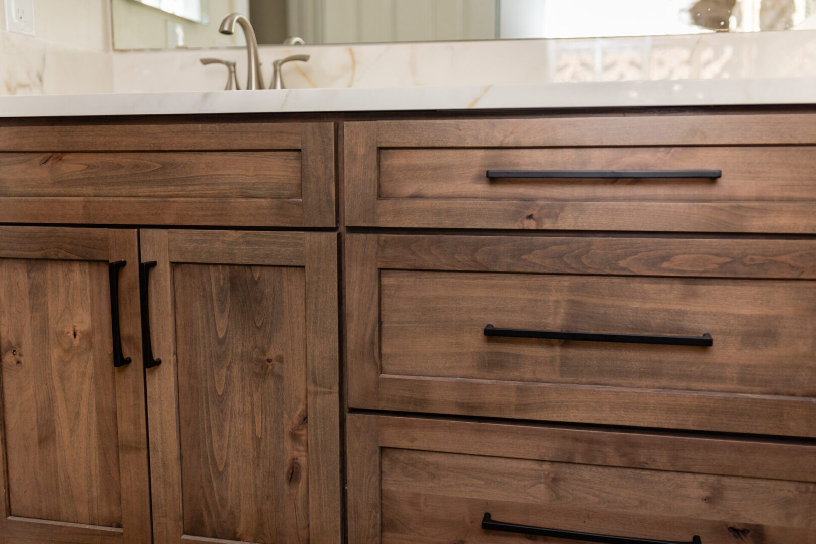 Wooden bathroom vanity with white countertop and sink faucet, featuring dark hardware and recessed panel cabinet doors