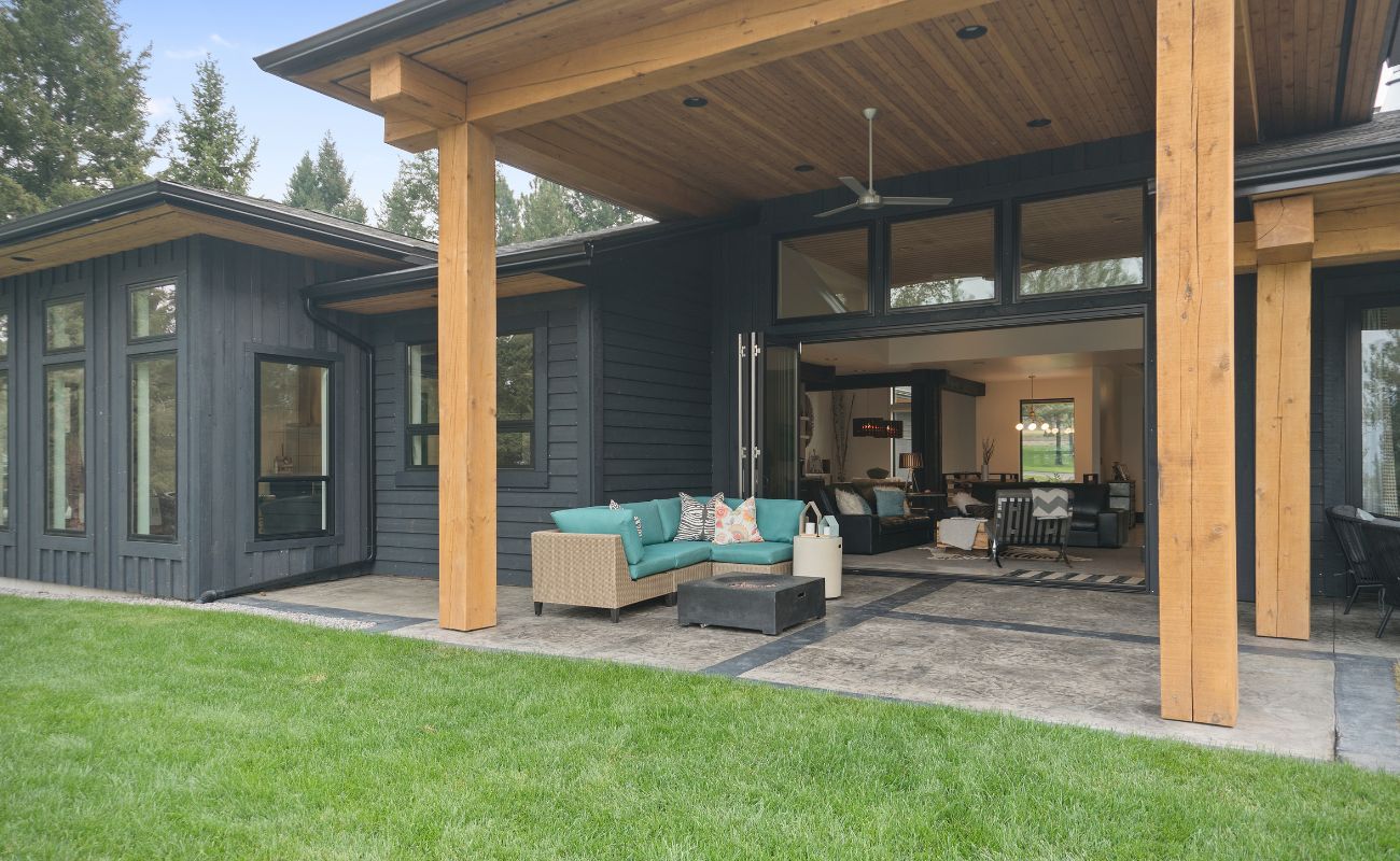 Modern home exterior with covered patio featuring turquoise sectional sofa, wooden beams, dark siding, and large glass sliding doors opening to interior living space