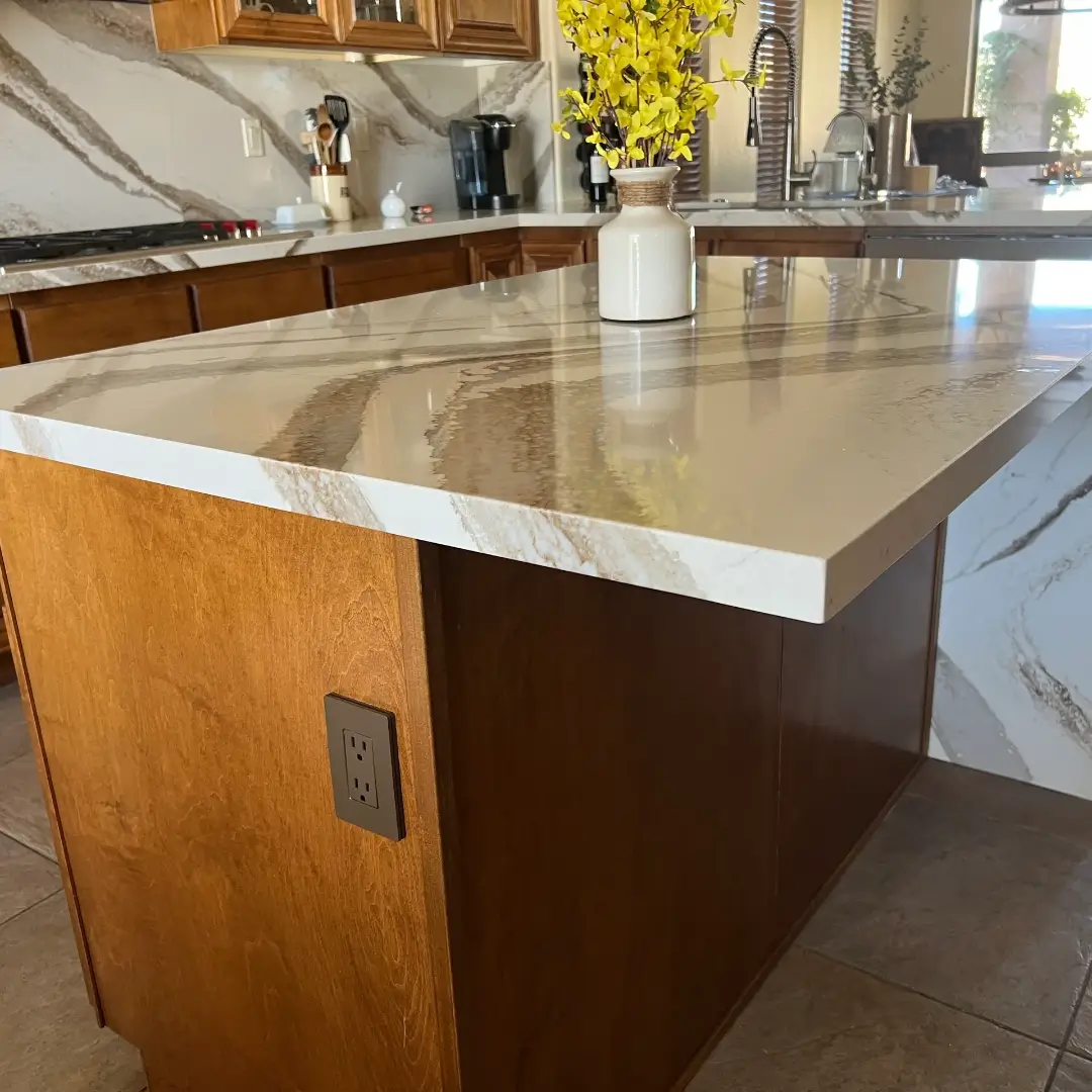 Modern kitchen with white and gray marble countertops, wood cabinetry, island with outlet, and yellow flowers in vase