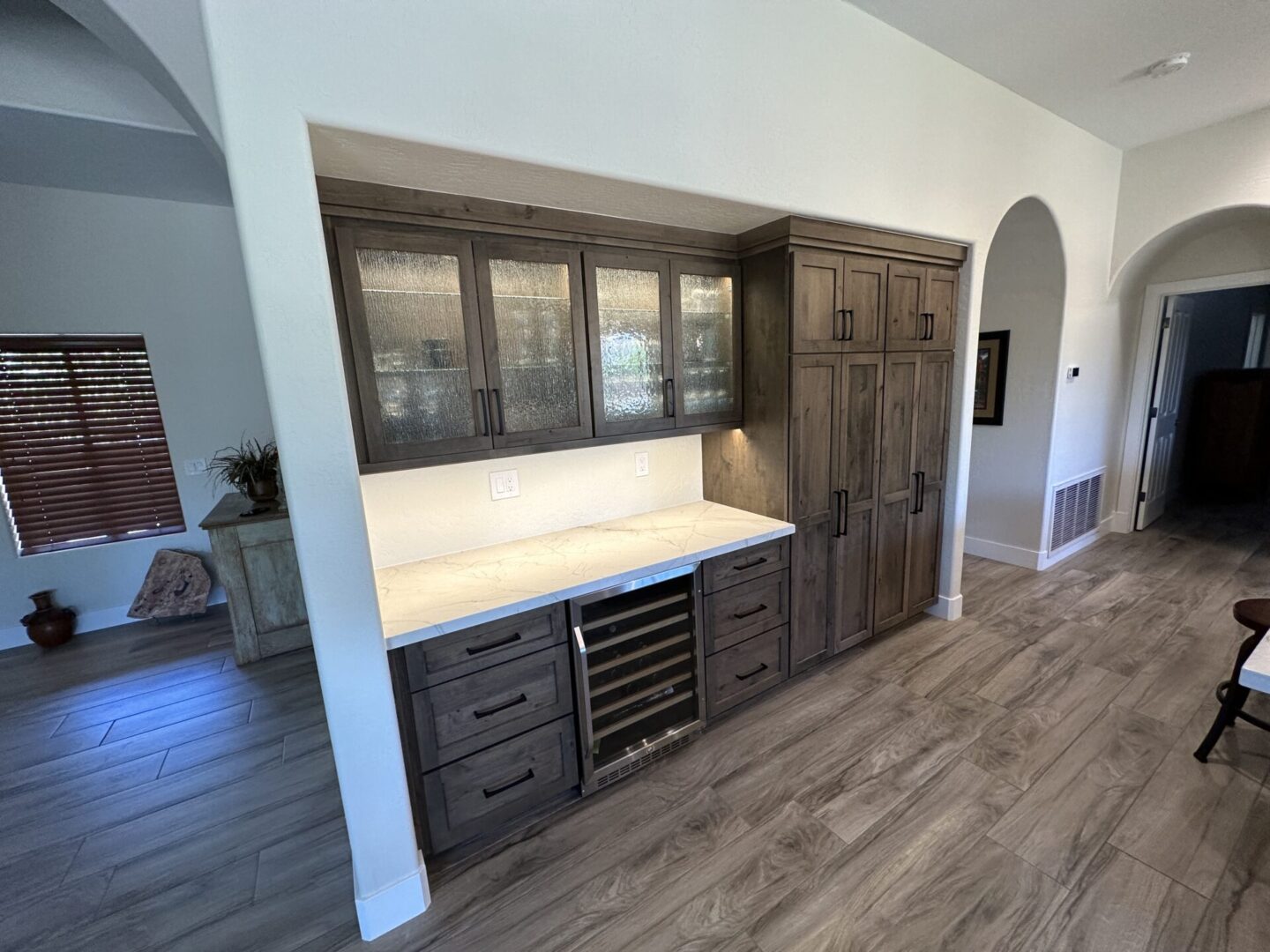 Modern home bar with dark wood cabinetry, glass-front upper cabinets, light marble countertop, built-in wine cooler, and tall storage cabinets against white walls with wood-look flooring