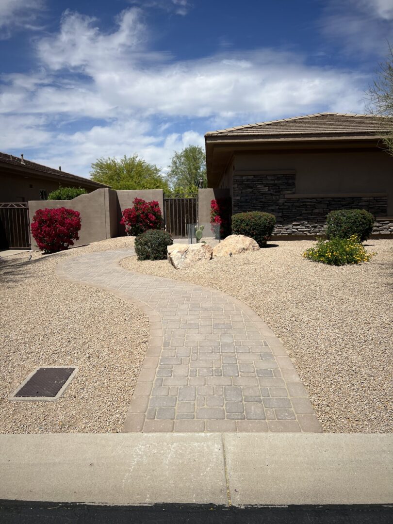 Modern desert landscaping with paved pathway, gravel, vibrant red flowering shrubs, and contemporary home with stone accent wall