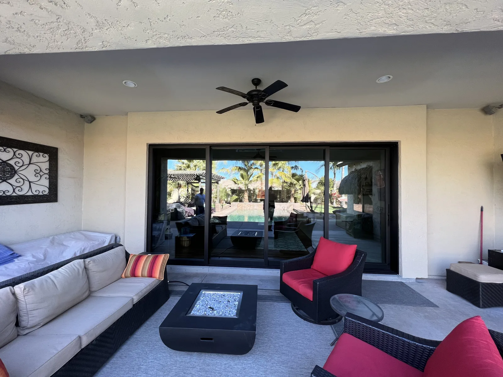 Modern patio lounge with black frame sliding glass doors, cream and beige furnishings, red accent pillows, black fire pit table, ceiling fan, and view of palm trees and pool beyond
