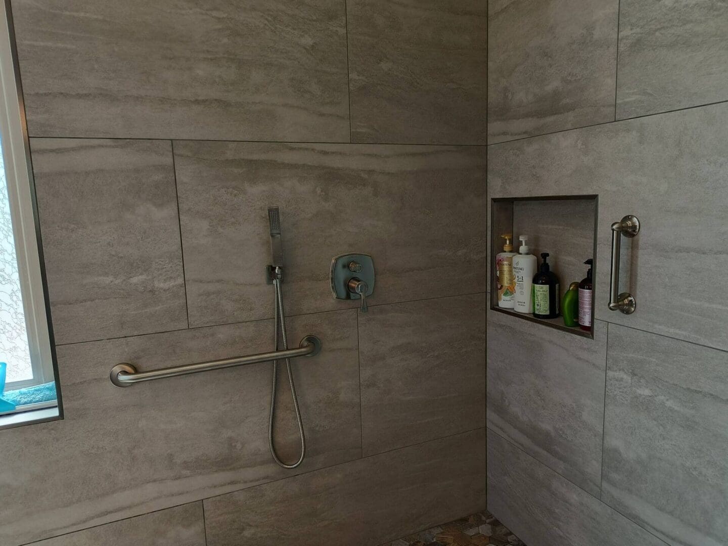 Modern accessible bathroom shower with gray tile walls, handrails, adjustable showerhead, built-in shelving with toiletries, and grab bars for safety and accessibility
