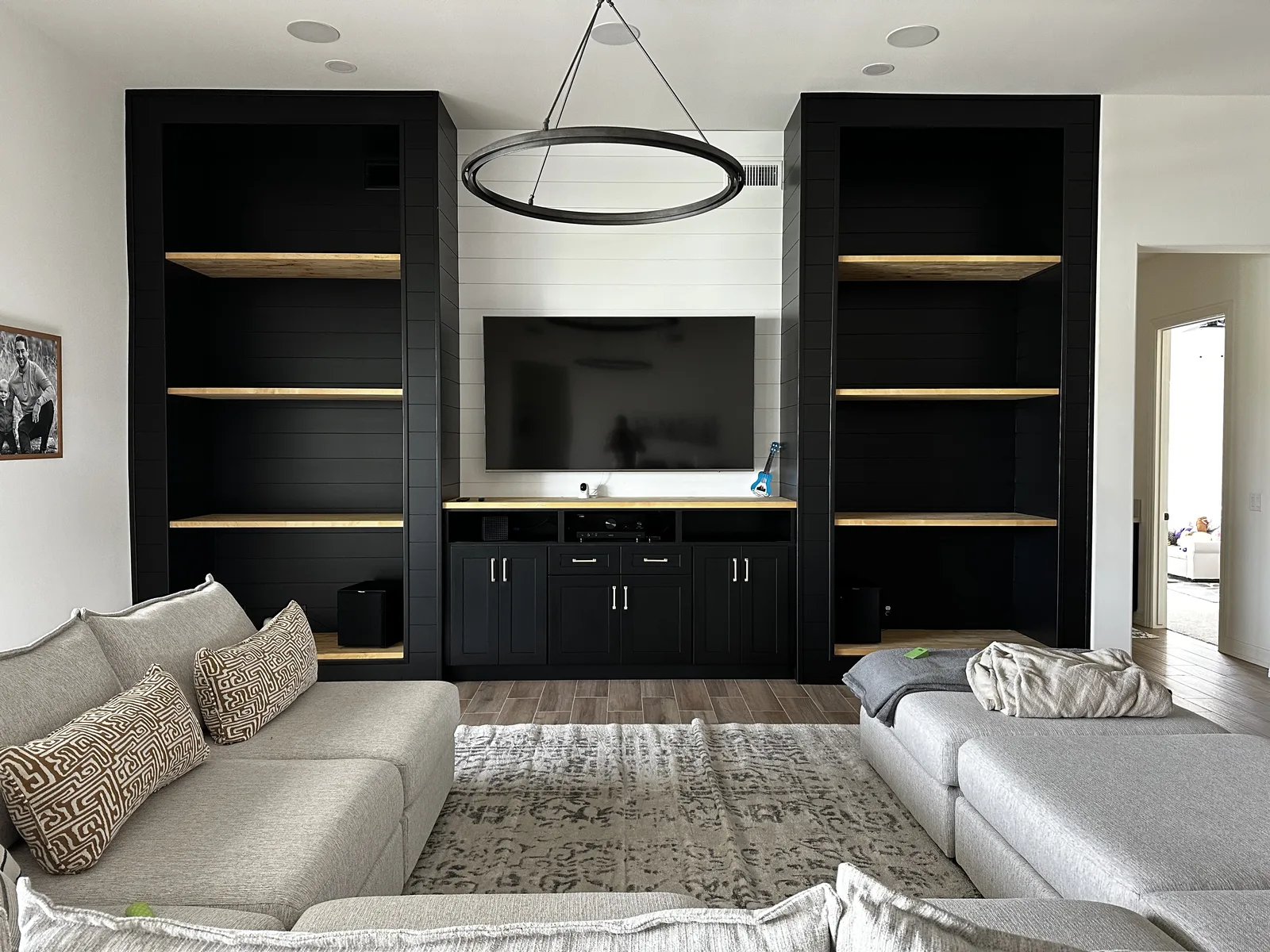 Modern living room with black built-in shelving units flanking a wall-mounted TV, wooden shelf accents, gray sectional sofas, pendant light fixture, and white shiplap accent wall